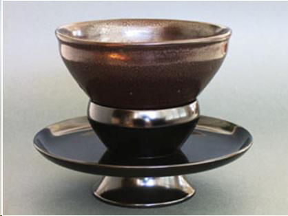 An "oil spot" temmoku bowl on a cup stand. The bowl is conical-shaped with a small foot. There is an indentation near the rim. The stand is wood. The bottom section of the stand is like a cylindrical ring flaring at the bottom. On top of the ring a shallow saucer about seven inches across is attached. A cup shape is attached on top of the saucer. It is about 3 1/2 inches across at the top. The foot of the bowl sits in this cup. In China, a stand like this was used to present wine cups and tea cups to people of rank. The idea may have been to show respect by preventing the servants from touching the cups of the more important people.