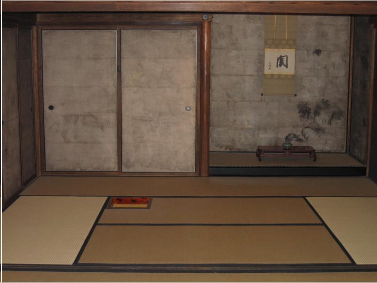 An eight tatami mat tea room. On the right is an alcove covered with sliver-grey paper. A scroll with a Japanese character hangs in the alcove. There is a low Chinese-style table with a celadon vase full of flowers in the alcove. On the left two sliding doors lead to a closet or another room. They are also covered with silver-grey paper. A small hearth is cut in the floor. The edge of the hearth is lined with a red and black lacquer frame.