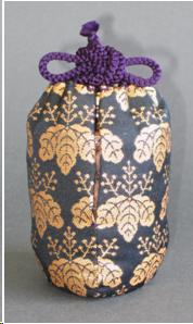 A blue brocade bag with a gold design of paulownia leaves. It is cylindrical in shape. The purple cords of its drawstring are tied in a bow knot.