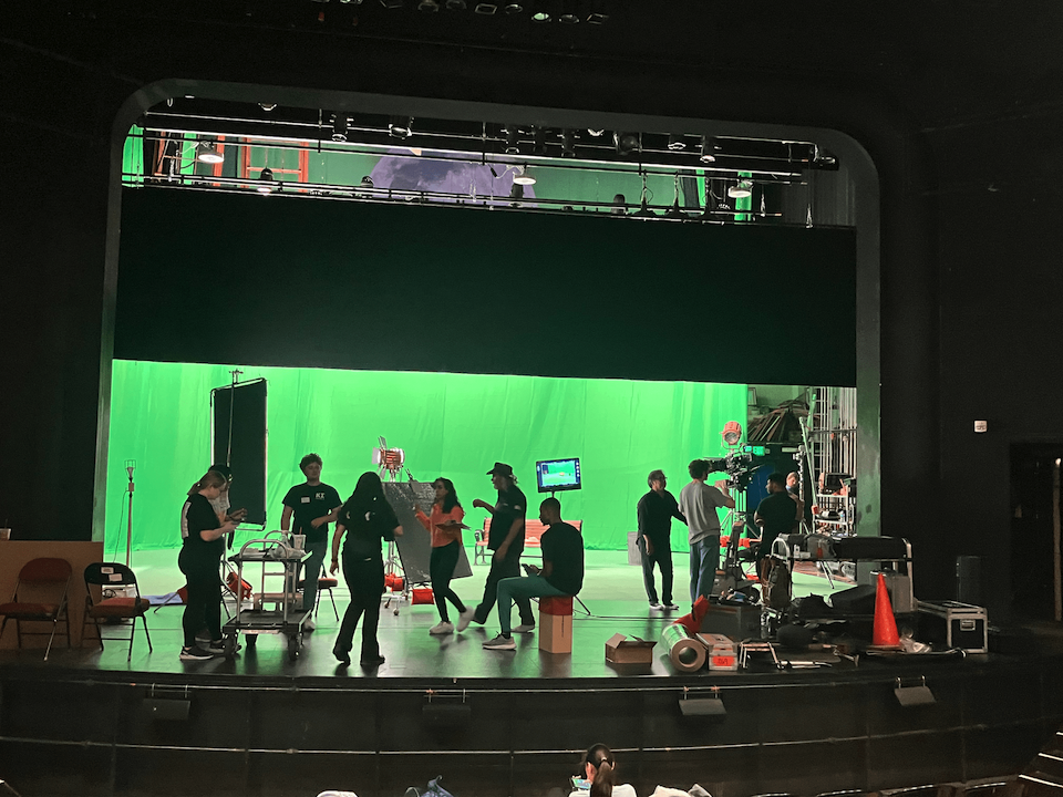 stage with green screen and multiple cast and crew milling about