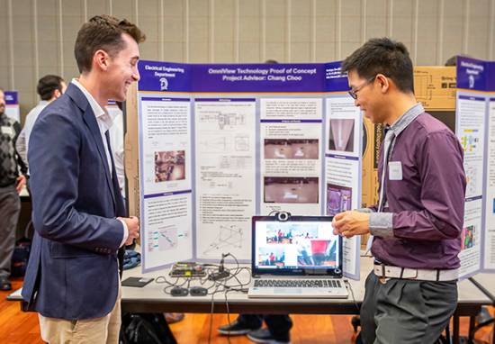 person stands in front of poster presentation with electronics on table talking and chuckling with guest
