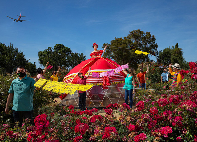 A live art installation of a mexican style tent dress sits in the middle of a rose garden and surrounded my men and women in traditional mexican clothing holding papel picado.