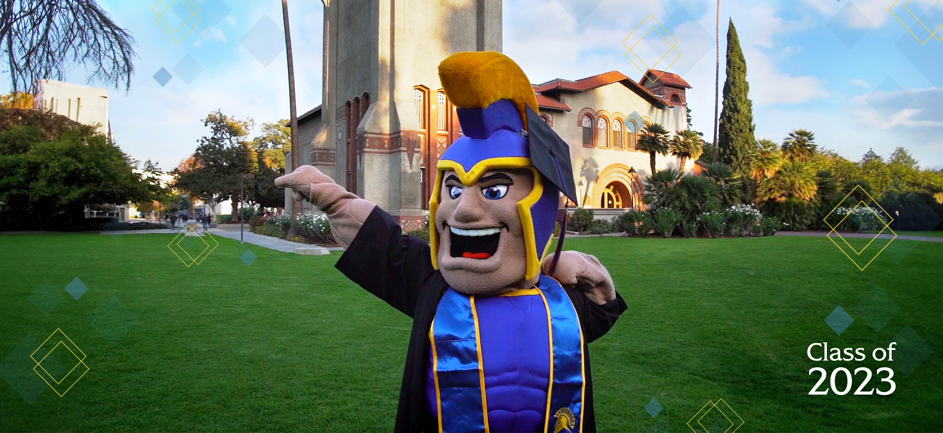 Sammy in grad gear posing with one arm flexed and the other extended out in celebration in front of Tower Hall.