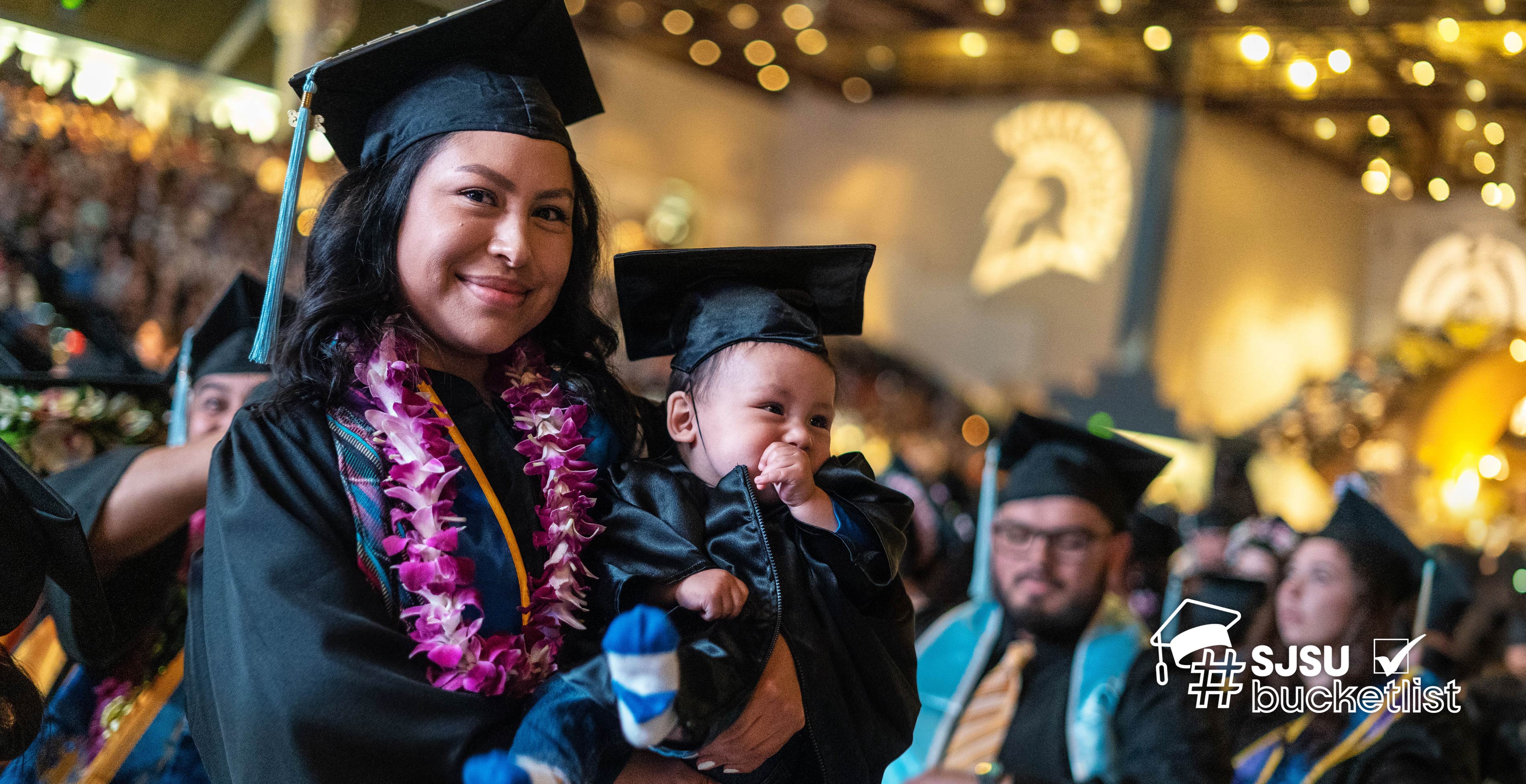 A graduate holding her baby as they giggle under the shimmering gold lights of the event center and awaiting graduates in the background.