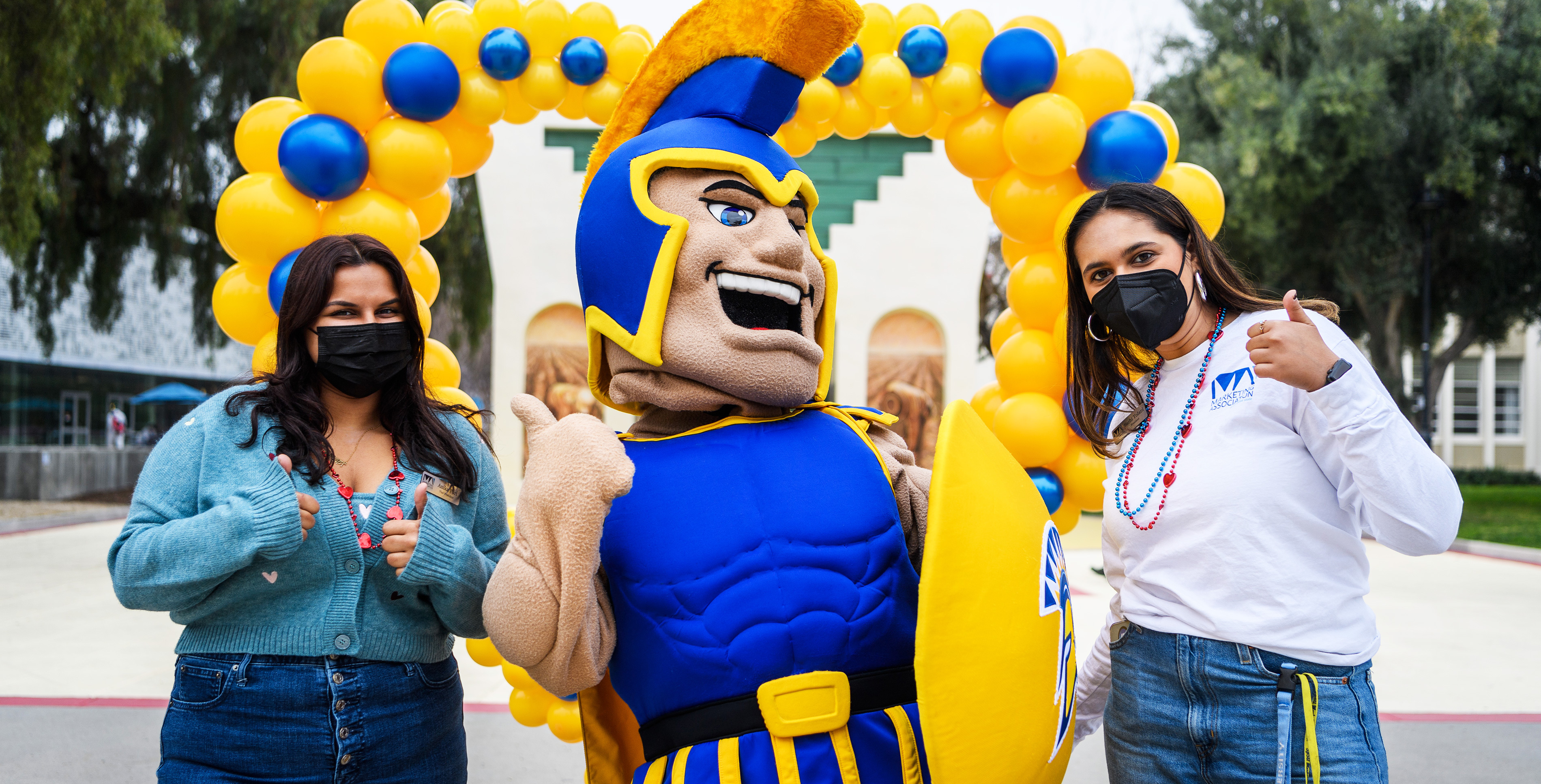 Two students with Sammy Spartan in the middle giving a thumbs up with a gold and blue heart shaped balloon display in the background.