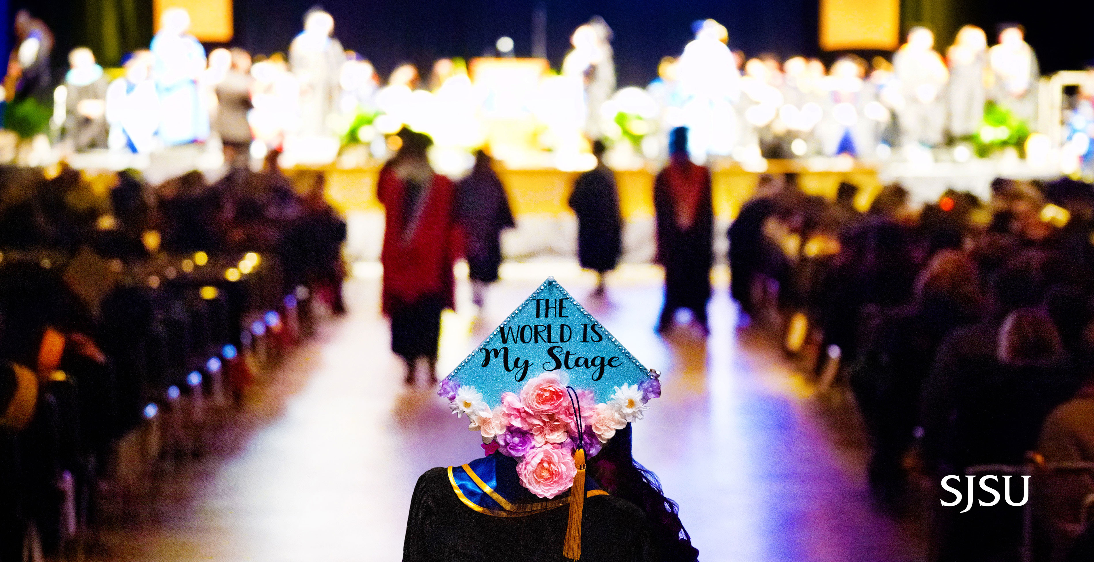 A single graduate stands in front of the pathway leading up to a lit up stage, her decorated grad cap shimmers with the words The World is my Stage.