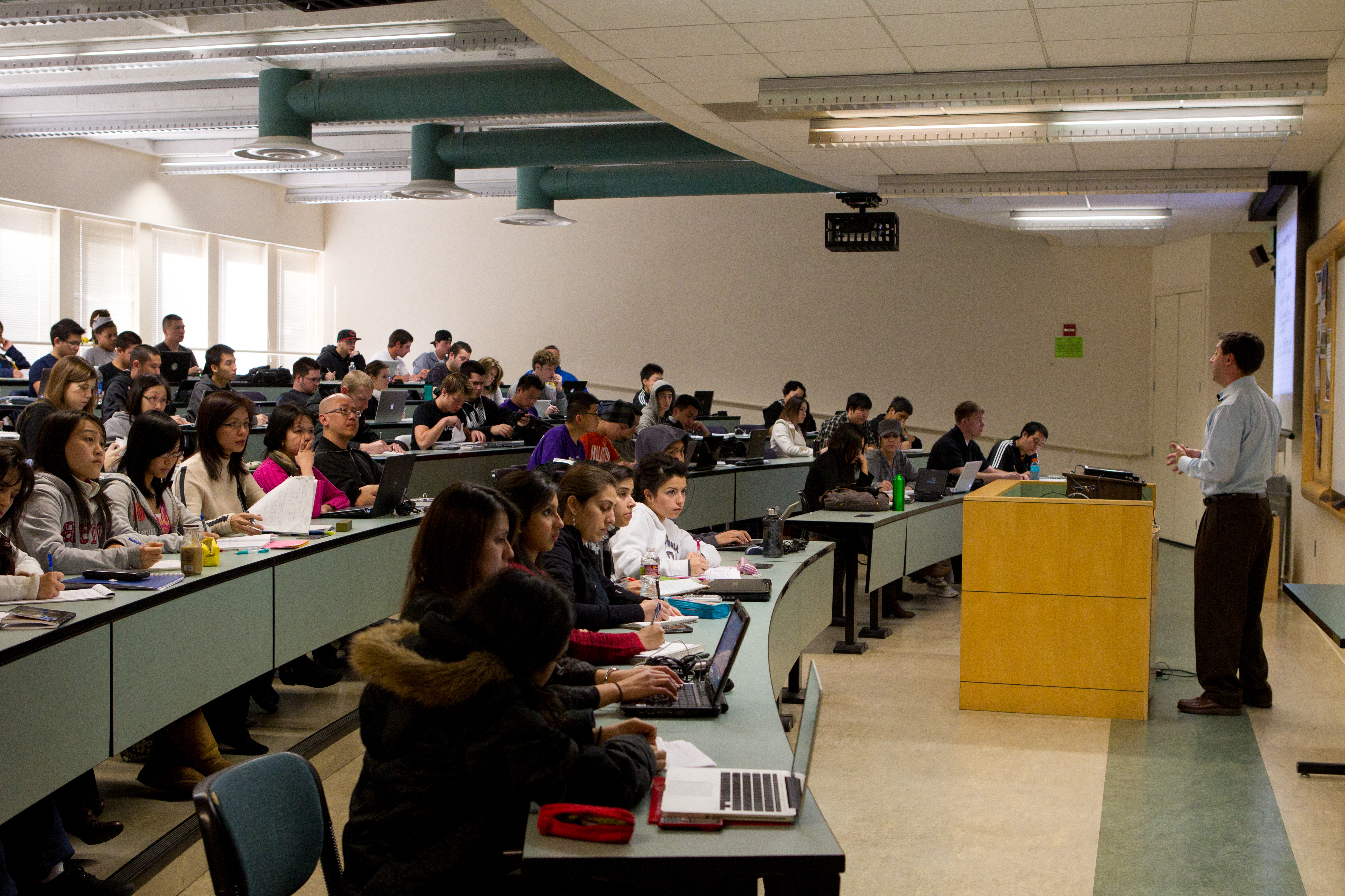 A large lecture hall with students taking notes on laptops paying attention to their expert professor.