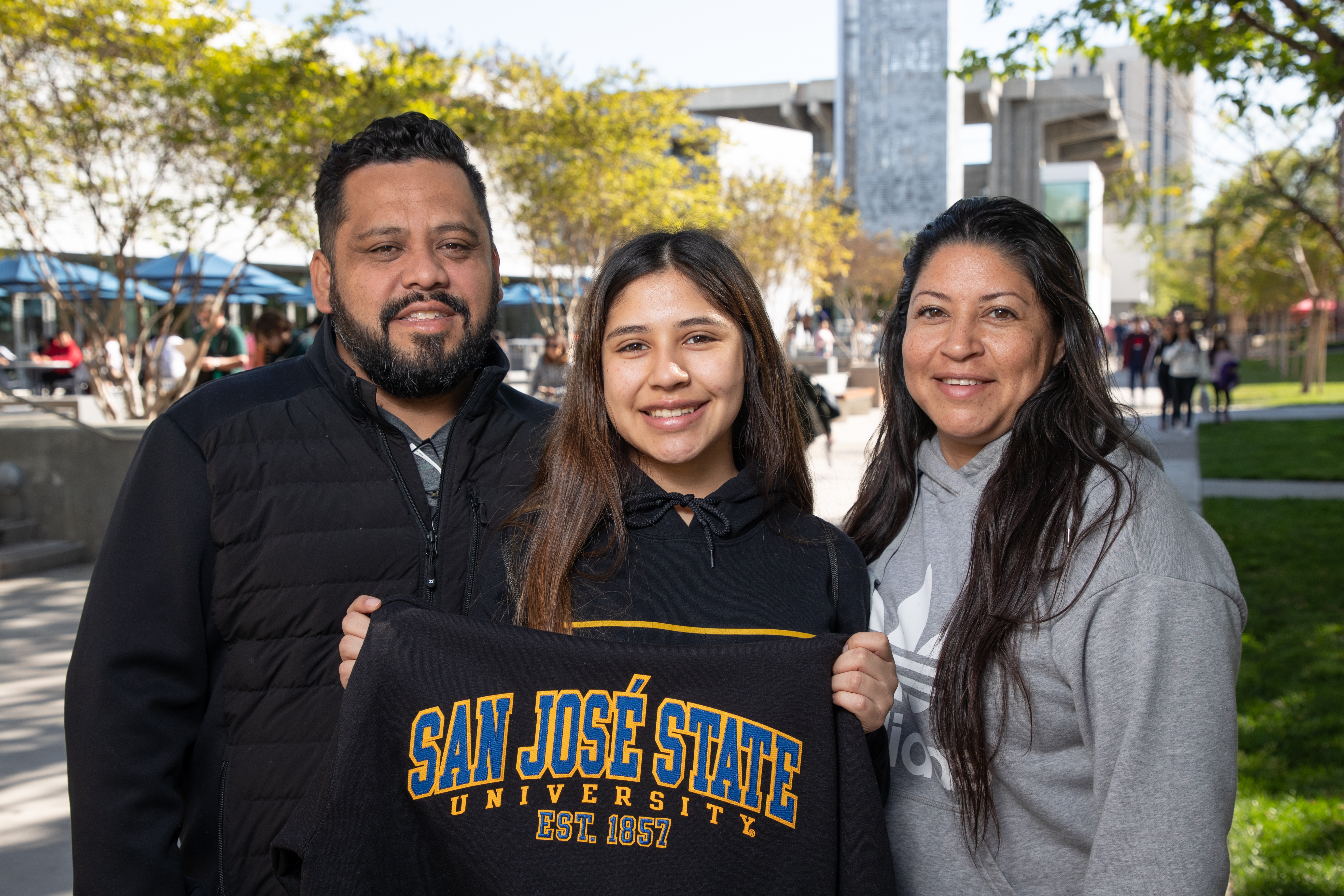 A student stands outside on campus with parents holding a SJSU sweatshirt.