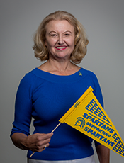waist-up photo of Marcia Daszko, Caucasian female with medium length blonde hair, wearing blue long sleeved blouse with sleeves pushed up to ellbows, holding a yellow and blue SJSU Spartan flag