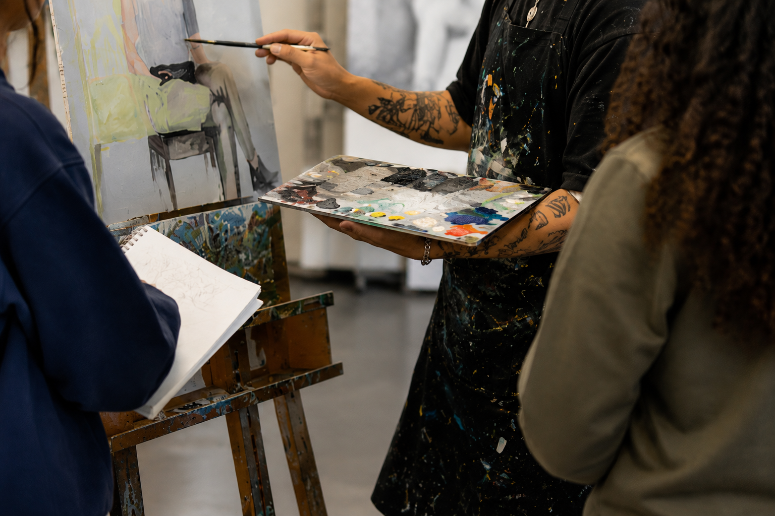Close-up of an MFA student demonstrating painting techniques at an easel while undergraduate students observe and sketch, with faces cropped out of the frame.