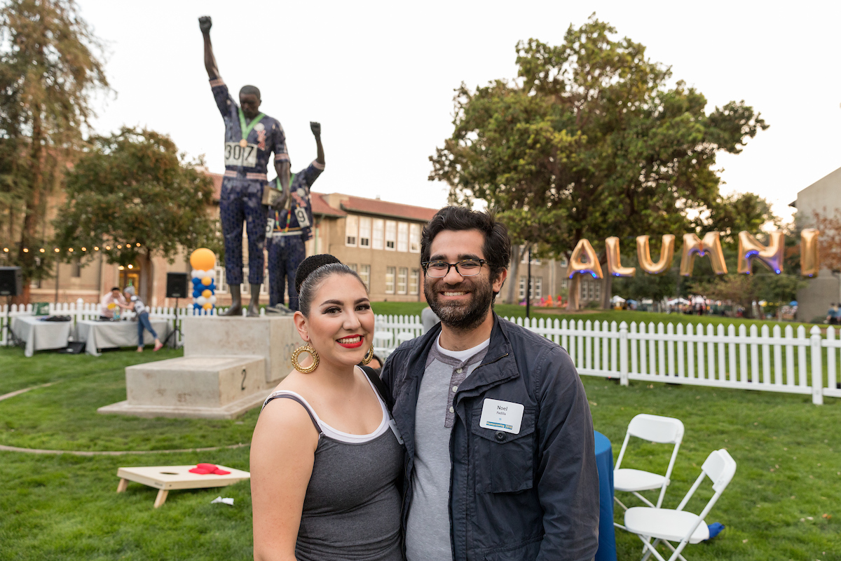 Two alumni pose in front of the Smith and Carlos statues at SJSU.