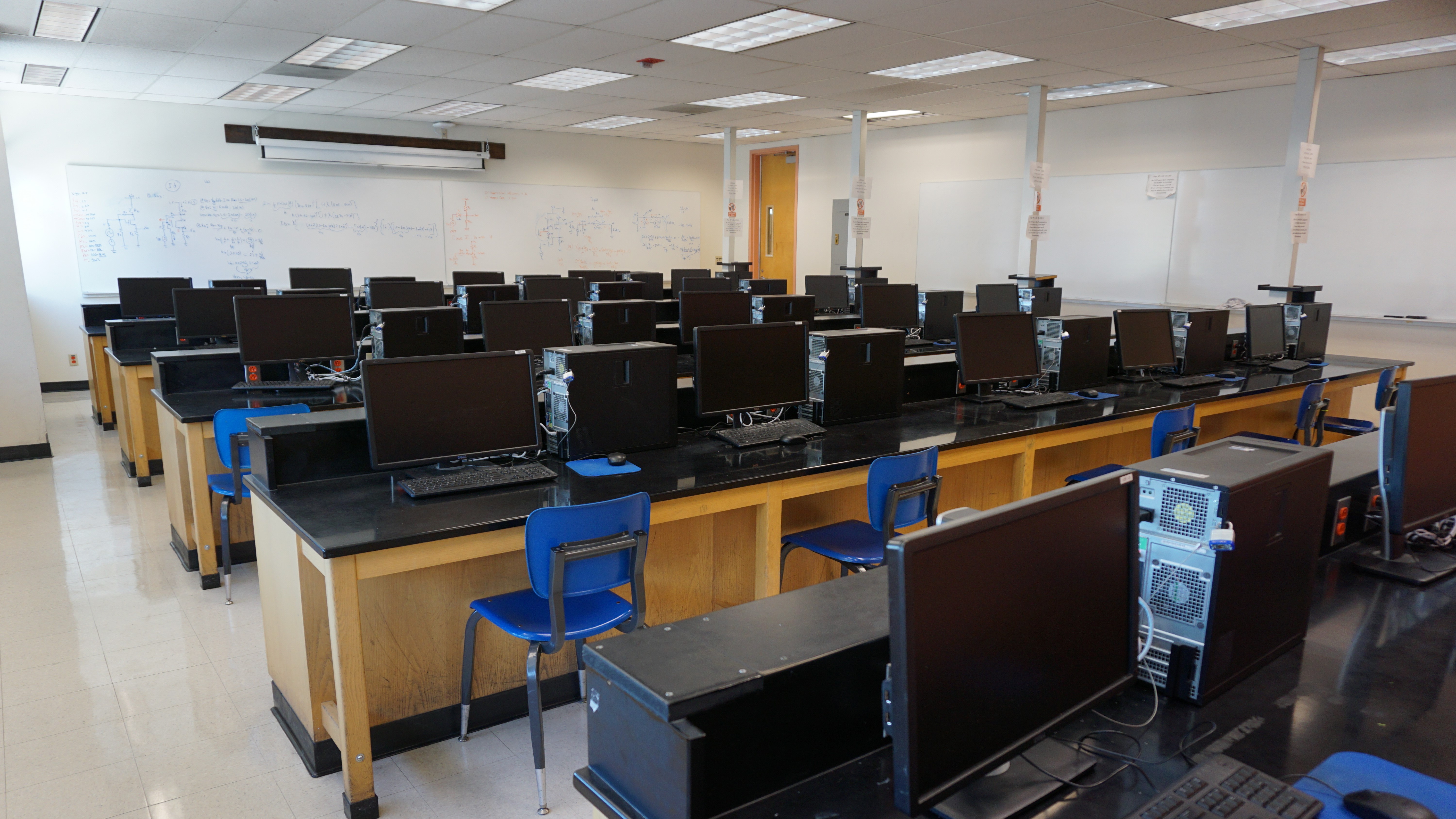 Black computer stations filling up a classsroom. Blue chairs are set up in front of every monitor.