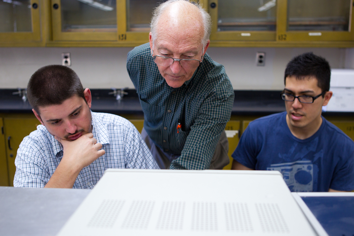 A professor stands between two students and helps them with an assignment.