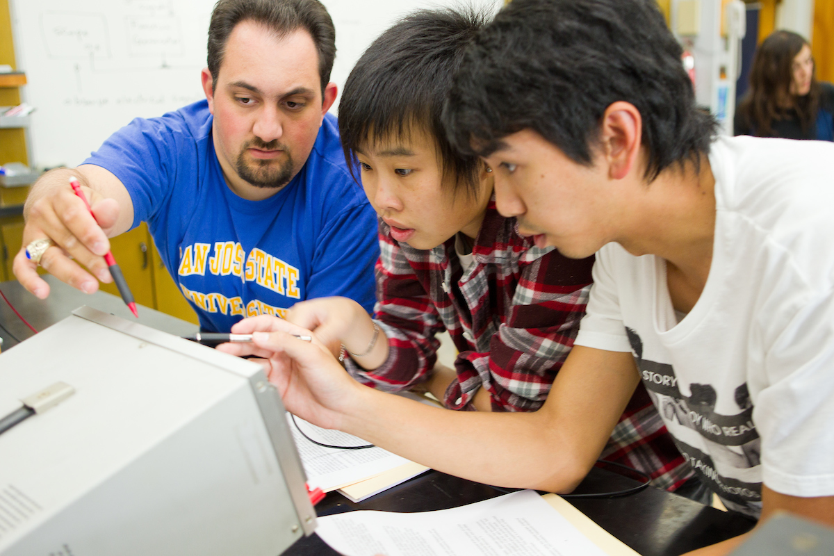 Three students gather around a monitor and point to it with their pencils.