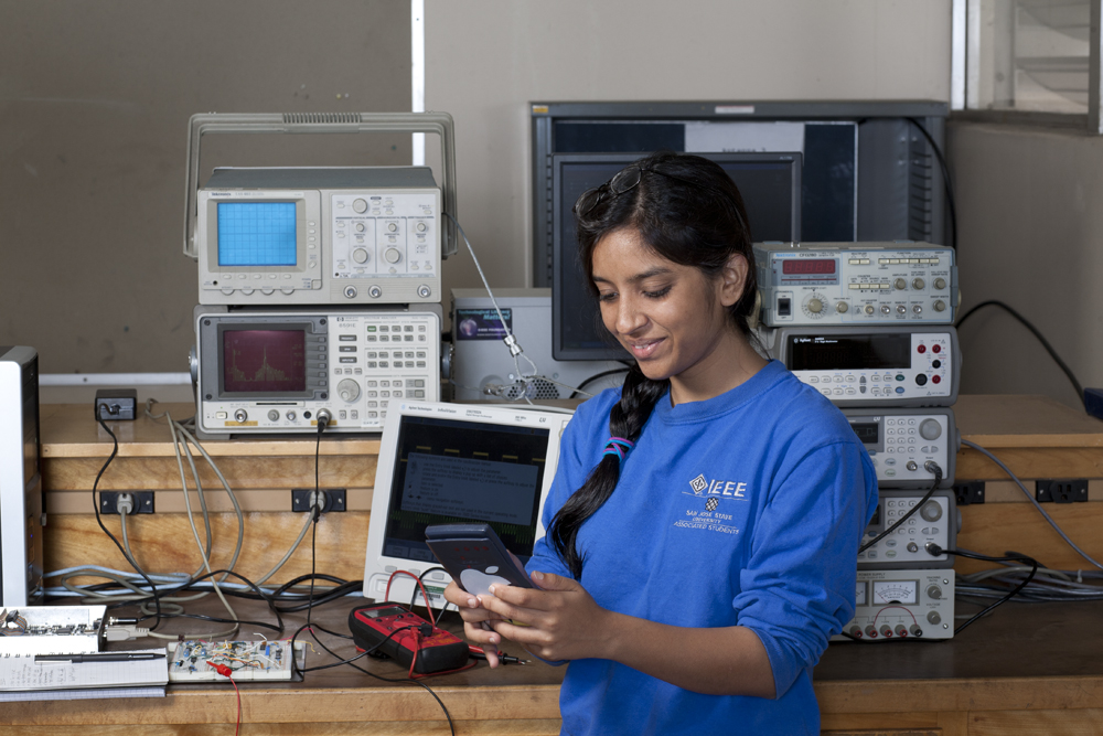 A student in a blue shirt is in a lab with many electronic devices behind her.
