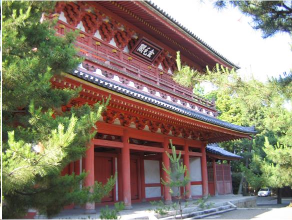 An elaborate white and vermillion, two-story Chinese-style gate. The roof is tile and the eaves are elaborately detailed. There is a sign with Chinese characters on it over the gate. Red pillars support the eaves on the first level. It is called a gate but it is really more like a building. There are pine trees on either side of the entrance.