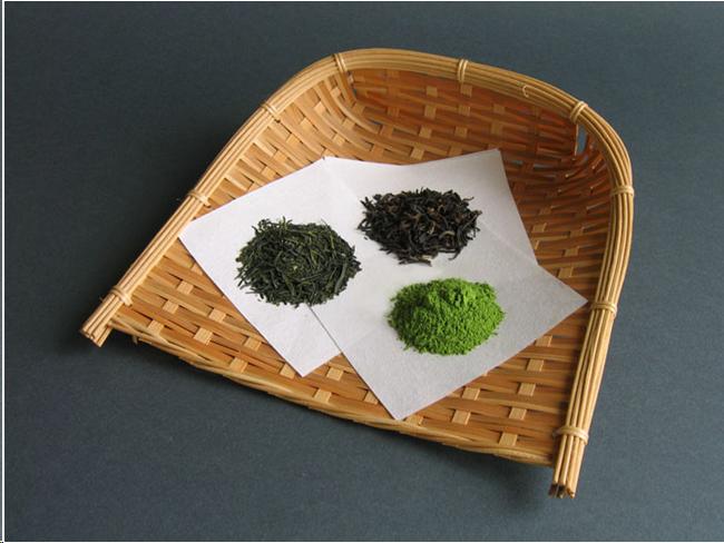 A straw tray like those used to winnow rice holds three white sheets of paper. On each sheet is a pile of tea. One pile is dark and coarse. It is roasted tea. Another pile is greener. It consists of rolled leaves which have not been roasted. The final pile is vibrant green. It is powdered tea.