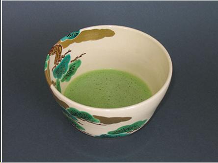 A cream colored bowl with a design of pine trees and gold clouds. The tea in the bowl is light green and the surface of the tea is covered with fine bubbles.