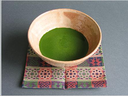 A buff and cream colored tea bowl sits on a square of brocade with green and purple designs woven into it. The tea is the bowl is grass green and has no bubbles.