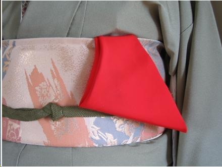 The mid-section of a woman wearing kimono is shown. Her kimono is light blue and her obi is white with pink, blue, and gold designs. A blue cord encircles the obi. It is knotted in front. A red silk cloth is tucked in the top of her obi. It hangs down in a triangular shape.