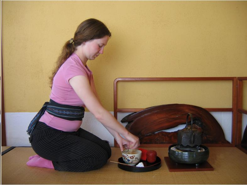A young girl with long red hair sits on her knees whisking tea. She wears a pink shirt and matching socks with black pants. In front of her is a tray with a tea bowl and various tea utensils. On the far side of the tray, an iron water pot sits on a small green ceramic brazier. Her lesson takes place in a tea room with tatami mats. 