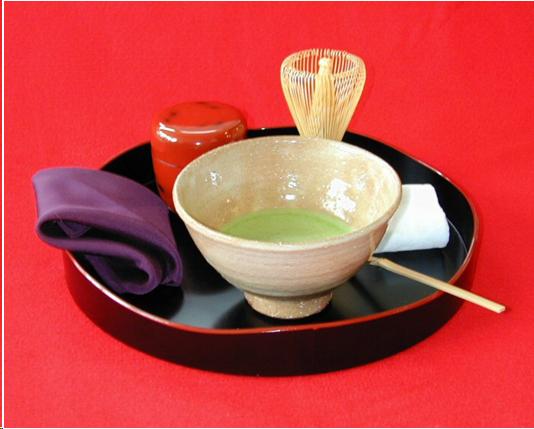 A black tray with an undulating red rim holds a tea bowl with frothy green tea, a tea whisk, a red lacquer tea container, a bamboo tea scoop, a small linen cloth for wiping the tea bowl, and a folded purple silk cloth for purifying utensils.