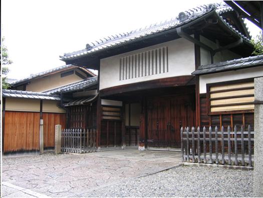 A two-story Japanese house gate. It has grey roof tile. The upper level is plastered white. There are slits in the wall so the people in the room over the gate can see who is outside. The gate doors on the lower level are wood with heavy iron hardware. The walls on either side are also roofed with tile. They are also made of wood and plaster.