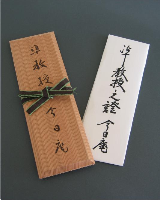 A tea certificate with its holder. The holder is made of two pieces of bare paulownia wood with Japanese writing done vertically on the surface in ink. The case has a brown ribbon with a green edge tied around the center. The certificate itself is inside a white, rectangular  envelope made of folded Japanese paper. It has black Japanese script written vertically upon it. The paper certificate can be sandwiched between the two wood boards of the case for preservation.