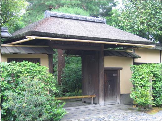 A one story Japanese gate with a thatched roof. The gates are open and we can see a garden inside. A low bamboo pole prevents people from walking in. The gate is set in a cream colored plastered wall. There is a small door in the wall to the right of the main entrance. 