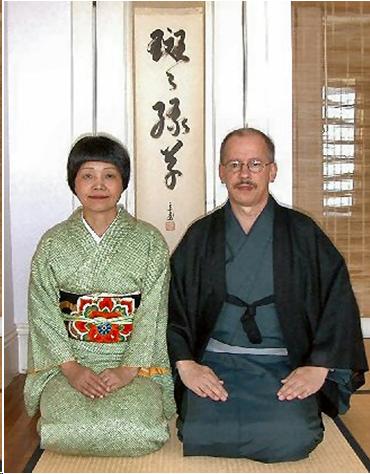 A Caucasian man wearing a blue kimono and a black kimono jacket sits next to a Japanese woman in a light green kimono and a colorful obi. They are sitting on a tatami mat in front of a Japanese scroll. He has a mustache and wears glasses. She has short black hair and smiles slightly. They are teachers of tea ritual.