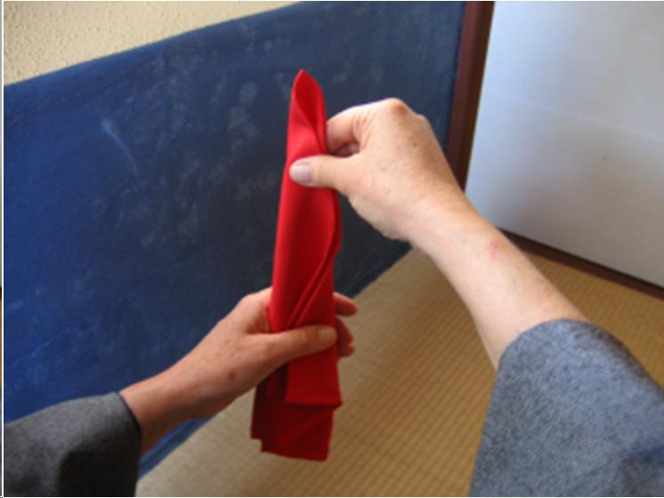 A picture of the hands of a woman wearing a blue kimono folding a red silk cloth used in the tea ceremony. She is sitting on tatami mats in a tea room. The cloth is suspended in a narrow triangle shape from the right hand. Her left hand assists.