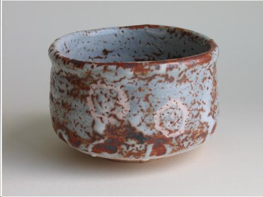 A grey and rust colored cylindrical tea bowl in the style of the Shino kilns.. It has two white abstract designs on the front which are six-sided geometric forms. They are supposed to represent the crane and turtle, emblems of good fortune in Japan. The bowl is irregular