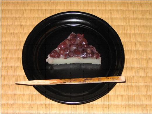 A round black lacquer dish sits on the tatami mat. It contains a triangular tea sweet. The top is covered with red adzuki beans. The bottom layer is a kind of sweet rice cake. A wooden pick used to eat the sweet rests across the front of the dish. 
