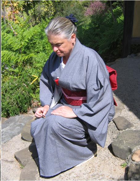 A woman in a blue kimono with a red obi squats down on the stone path to the tea house with one knee up. She holds her fan in her right hand and it rests on her right knee. Her left hand rests on her left thigh. Her eyes and head are lowered. 