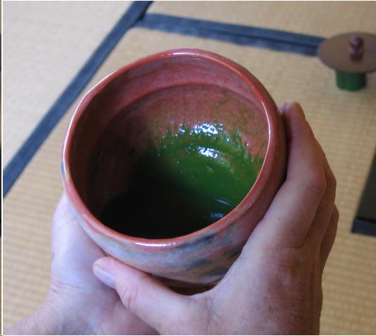 The guest at a tea ritual holds a red Raku-type bowl of thick green tea in her hands. Only her hands and the bowl can be seen. There are tatami mats and the bronze  lid of a tea kettle on a bamboo stand in the background.