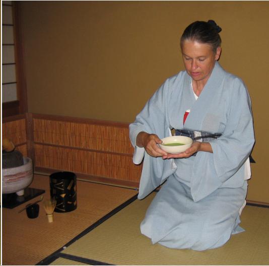 A woman in a light blue kimono and dark blue obi holds a shallow, white bowl of thin tea and prepared to drink. She is seated on tatami mats. To the viewer's left a black water jar with a gold design is seen. A black lacquer tea container and a bamboo whisk sit in front  of the cylindrical water container. To the left of the water container, the corner of a ceramic braizer and iron kettle can be seen. There is a short bamboo screen behind the area where the tea has been made.
