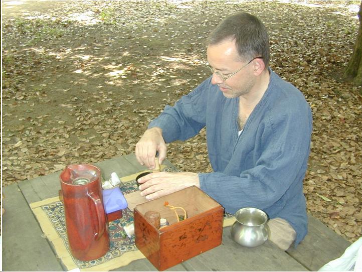 A man with short, dark hair and glasses sits at a picnic table in a park. He wears a blue, Japanese-style shirt. He is whisking tea. There is a wooden box for tea utensils to his left on the table. To the left of the box is a pewter bowl for waste water. A thermos bottle sits opposite the tea utensils the man is using. 