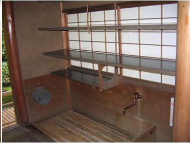 The area where tea utensils are washed and prepared for use in a tea house. The area consists of three shelves suspended over a bamboo grate which has a drain underneath it. The shelves are made of polished wood and there is a window covered by shoji screens behind the shelves. Underneath the lowest shelf, a copper faucet is located. The area is about six feet long and three feet deep. The lowe part of the alcove is lined with wood. The upper part of the walls are made of brown clay. 