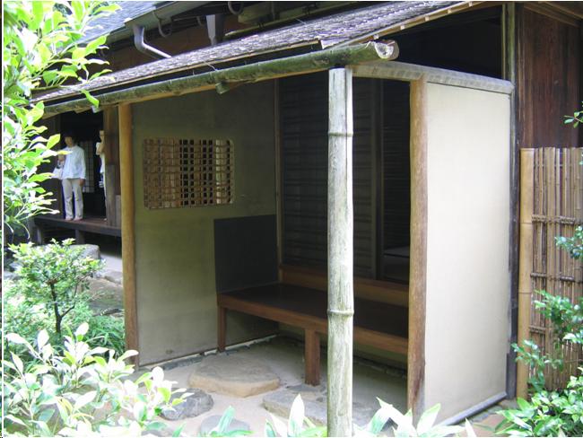 A small shed-like structure containing a wooden bench on which the guests sit as they wait for a tea gathering to start. The roof is covered with cedar shingles and the front edge is lined with a bamboo rain gutter. The waiting area is connected to a larger building. In the wall on the left side of the structure, there is an opening like a window with a bamboo grating across it. Stones are set in the cement in front of the bench on which the guests can rest their feet. Shrubs and bamboo can be seen in the tea garden in front of the waiting bench.