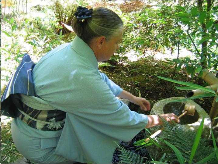 A women wearing a light blue kimono and a dark blue and silver obi squats down to purify her hands and mouth at a stone basin. She holds a bamboo dipper in her right hand. There are ferns and shrubs in the background.