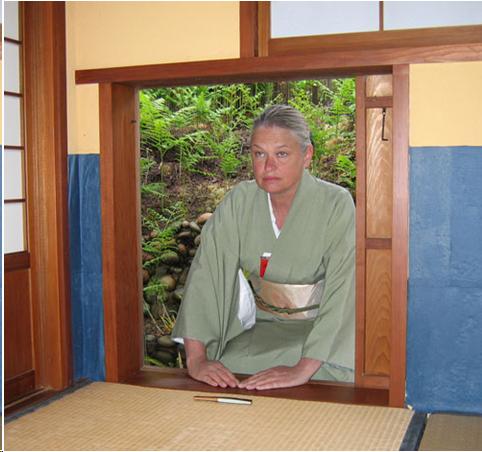 A woman in a light green kimono prepares to enter the "crawling in" entrance of a tea room. This small wooden sliding door is about three feet square. The woman's hands rest on the threshold and her fan is placed on the tatami mat just inside the room. The green ferns of a tea garden can be seen behind her. 