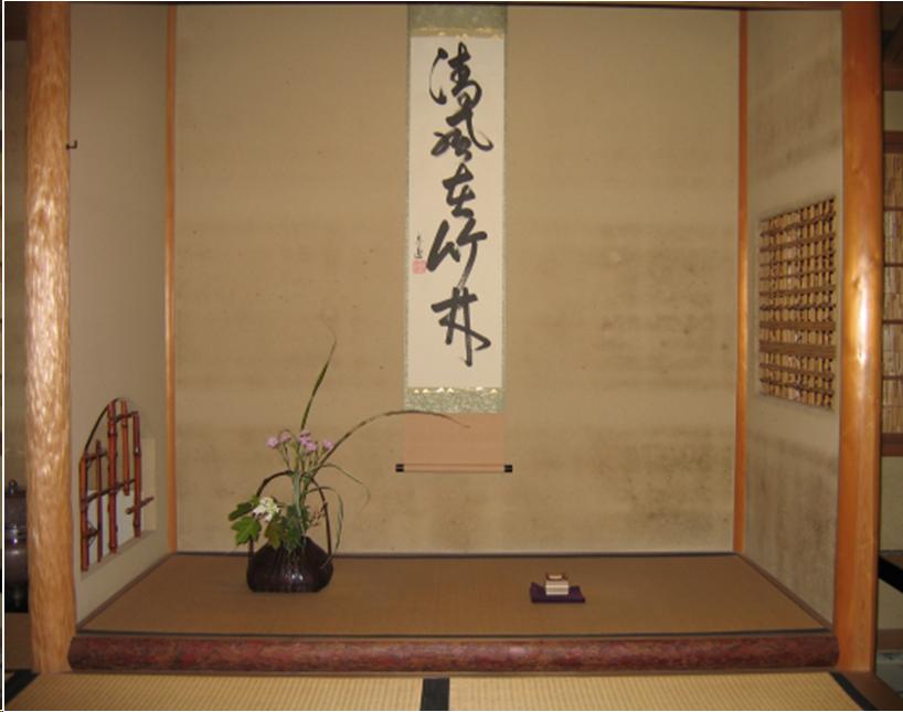 The alcove of a tearoom. It is six feet wide and three feet deep. On the left side the front vertical support is an irregular, blonde piece of polished wood from which the bark has been removed. The support on the right is more regular and also made of polished wood. A darker piece of wood with the bark on it lines the bottom of the slightly raised floor of the alcove. The floor is covered with a tatami mat. In the right wall of the alcove there is a square opeing with a bamboo grate. It is located about two and a half feet up the wall. In the left wall, there is a arched opening with another bamoo grate. There is a scroll with Japanese script on the back wall of the alcove and a basket of flowers sits on the floor of the alcove to the left of the scroll. An incense container on a purple cloth sit on the floor to the right of the flowers.