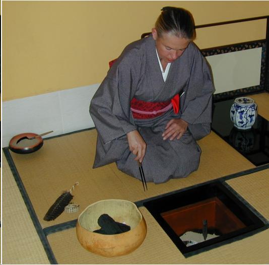 A woman in a dark blue kimono and red obi sits on the tatami mat in front of a small square hearth cut in the floor of the tearoom. She hold metal chopsticks in her right hand as she prepares to place charcoal in the hearth. To the left of the hearth is a large gourd basket containing black wood charcoal. A feather and an incense container sit on the tatami behind the basket. There is a bowl of ash to the left, behind the woman. To the viewer's right is a blue and white water jar resting on a wooden board.