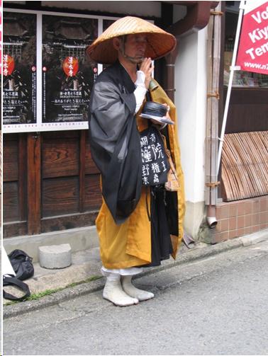 A monk with a straw rain hat stands on the street in Kyoto. One hand is raised in a gesture of prayer while the other holds his begging bowl. He wears a black kimono and with yellow robe over his shoulder. He stands on the street in white tabi socks.