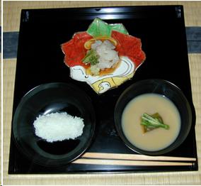 A black lacquer tray holding the first course of a tea ceremony meal. At the right front of the tray, there is a black lacquer bowl of white miso soup with some vegetables in the center. A cylindrical form of white rice in another black lacquer bowl is to the left of the soup. At the rear of the tray is a maple leaf shaped dish. It has a design of leavesin green, rust, and yellow. There is a pile of raw fish and some garnishes in the middle of the dish. 