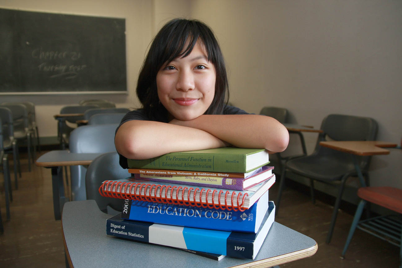 Black haired student looking at camera with a smile, leaning with both hands pressed on a generic stack of books