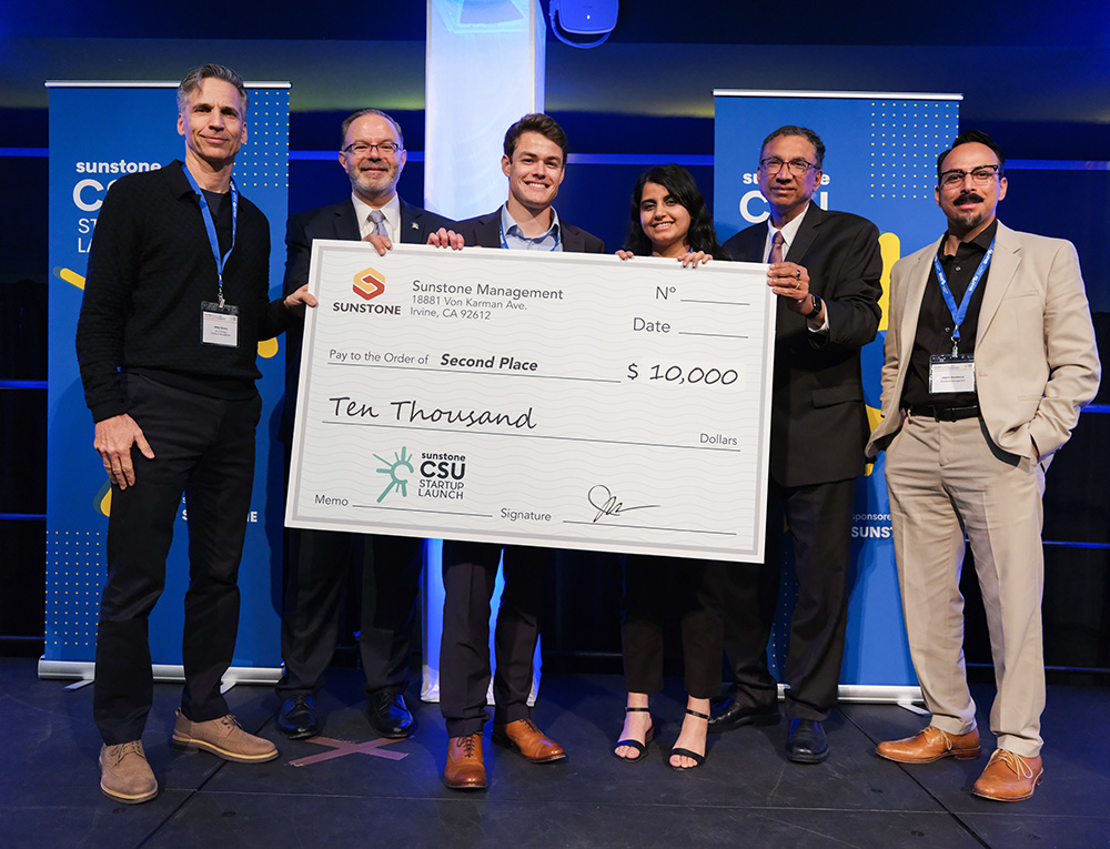 Two college-age startup founders pose onstage with two sponsors, two hosts, and a giant check for $10,000