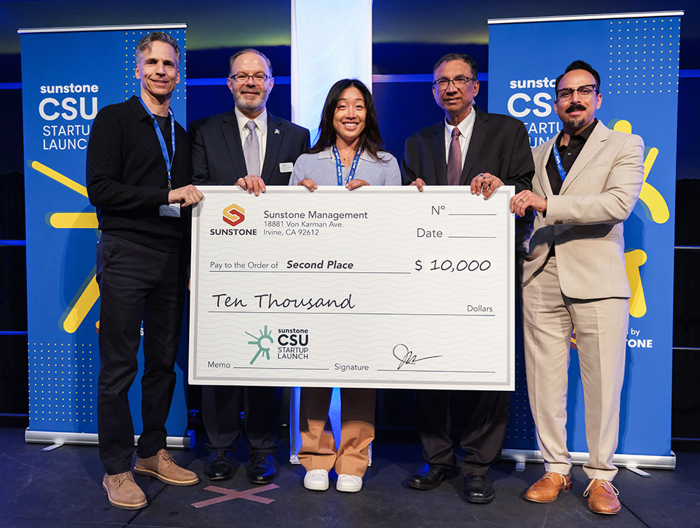 A female, college-age startup founder poses onstage with two sponsors, two hosts, and a giant check for $10,000