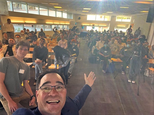 A dark haired man dressed in a sport coat and tshirt, laughing, takes a selfie with an amber-toned roomful of people behind him.