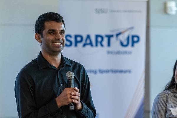 A dark-haired man holds a microphone and smiles, with a white pop-up banner that says "SpartUp Incubator" behind him.